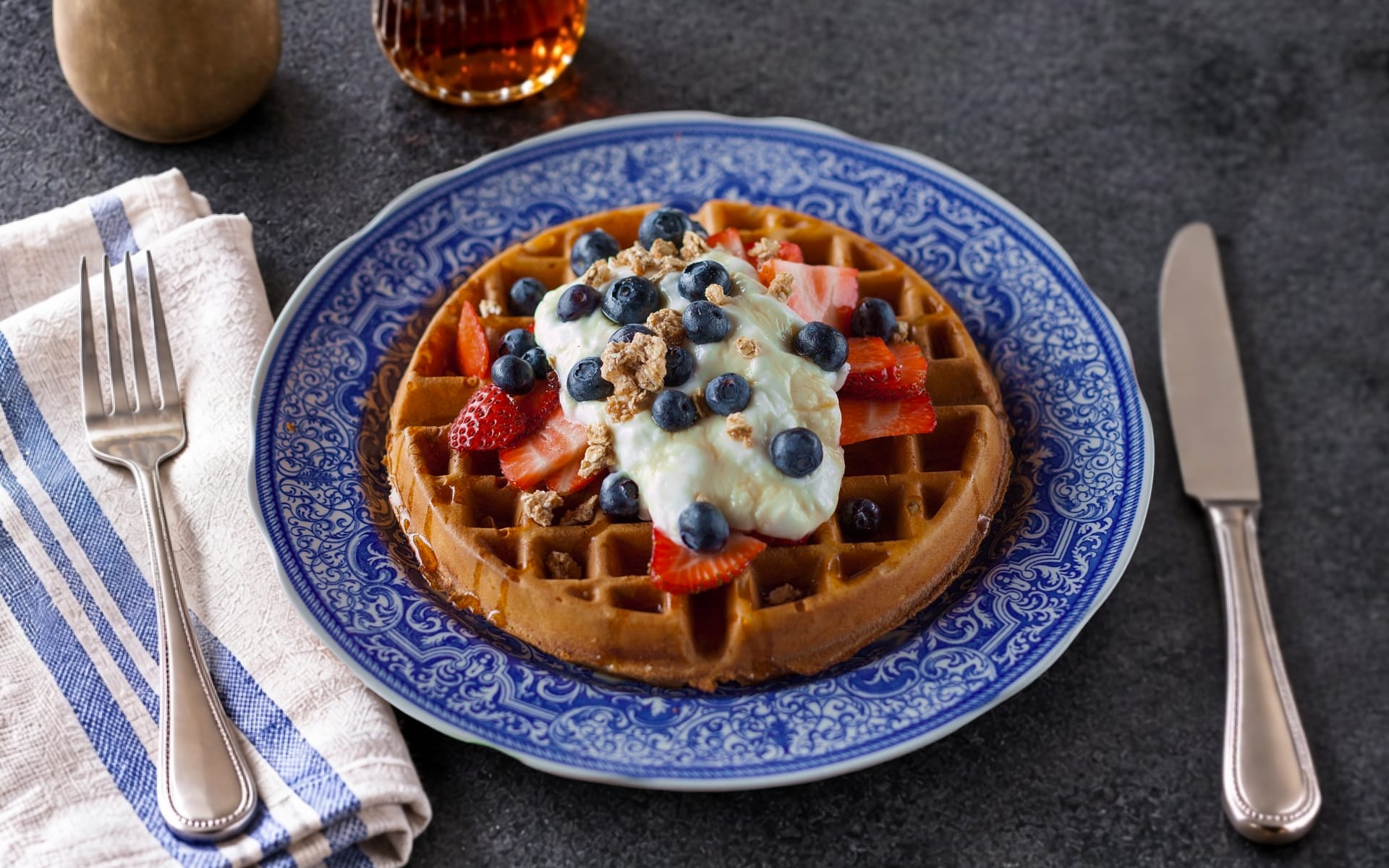 A freshly baked waffle on a blue patterned plate, topped with dairy-free yoghurt, sliced strawberries, fresh blueberries, and granola crumble.