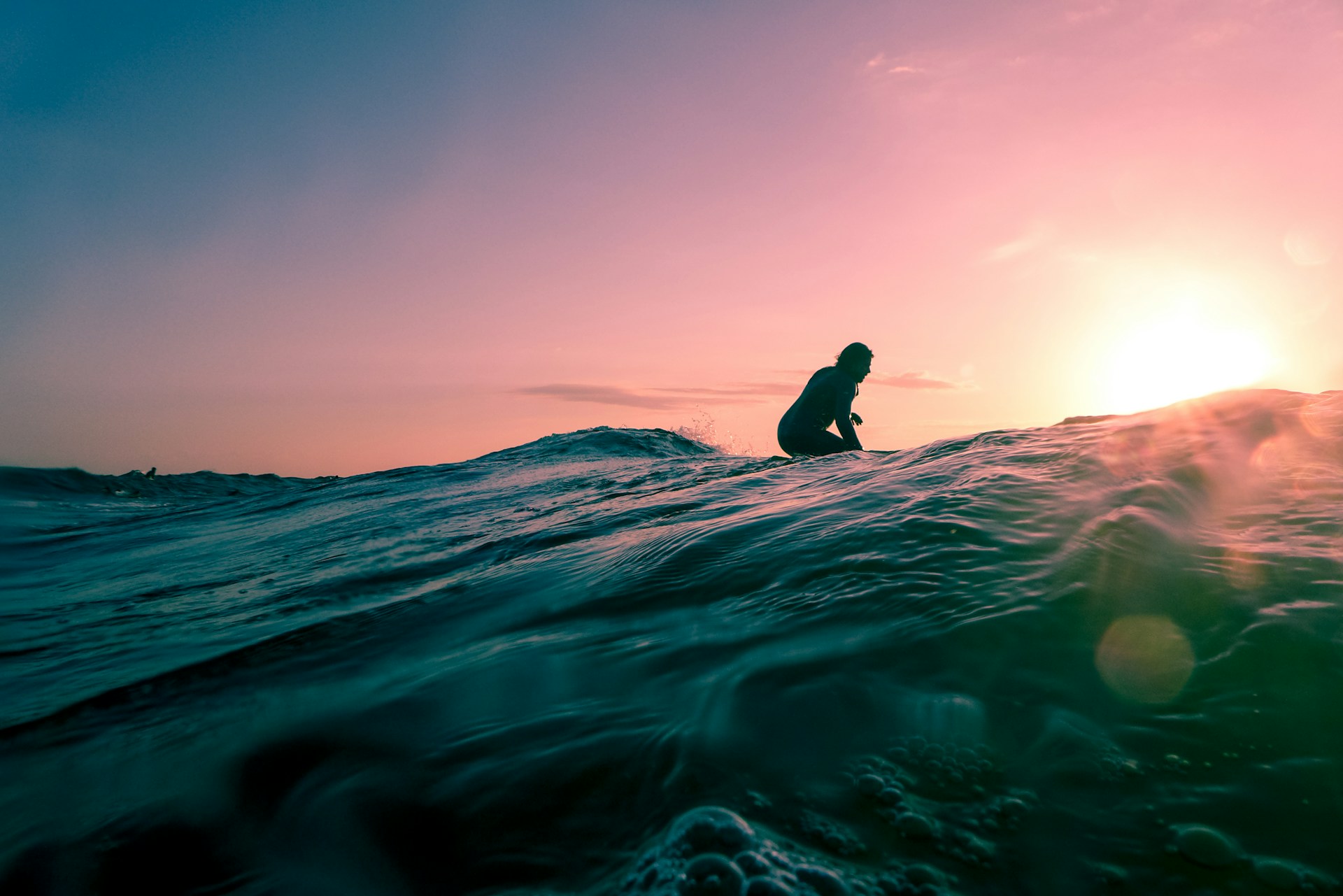 Silhouette of a surfer crouched on a board at the crest of a wave against a pink sunset, shot from water level